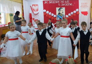 Dziewczynki w białych sukienkach z czerwonymi paskami oraz chłopcy ubrani na galowo tańczą w czwórkach Poloneza.
